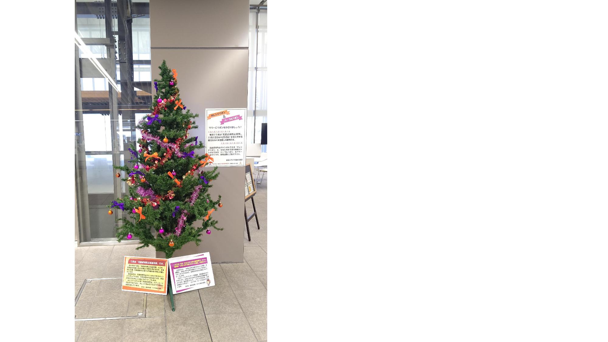 オレンジリボン・パープルリボンが飾り付けされたクリスマスツリーの下に、子どもへの虐待を防止運動と女性に対する暴力根絶運動の説明が書かれているボードが設置されている写真